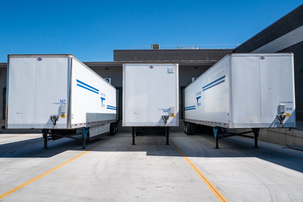 hero-img-01 Three white cargo trailers parked at an industrial shipping dock under clear blue skies.