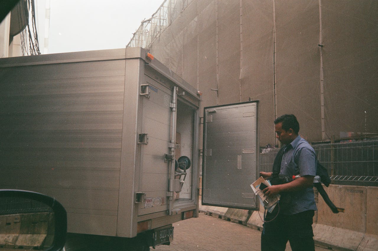 gallery-2 A logistics worker in a blue shirt checking a cargo truck on a city street. Industrial background suggests transportation and delivery.